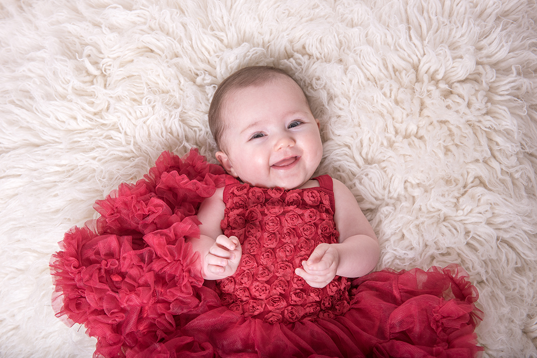 baby smiling in red dress laying on rug during norfolk baby photo session